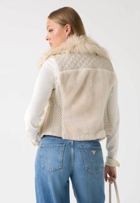 Gilet cropped beige con motivo trapuntato, colletto in pelliccia sintetica e texture morbide. Abbinato a jeans in denim chiaro. Palette di colori neutri.