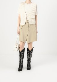 Chaleco acolchado beige con mangas cortas, blusa texturizada color crema, falda beige con bolsillos y botas vaqueras negras con bordado plateado. Bolso blanco.