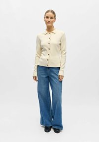 Femme portant un cardigan crème boutonné, un jean bleu à jambes larges et des chaussures noires, debout devant un fond clair uni.