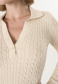 Maglione beige lavorato a maglia con scollatura a V, caratterizzato da un motivo a cavo testurizzato e un singolo bottone di chiusura al colletto. Materiale morbido e leggero.
