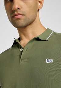 Olivgrön polo t-shirt med en strukturerad tyg, två knappar och en vit accentrand på kragen. Har en blå broderad "Lee"-logotyp.