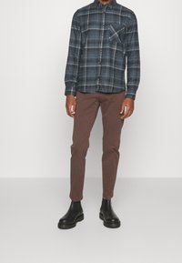 Geknoopte geruite shirt in donkerblauw en zwart, met een enkele borstzak, gecombineerd met bruine slim-fit broek en zwarte enkellaarsjes.