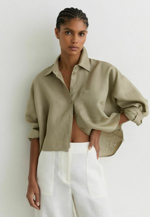 Žena s vrkočmi, ktorá má na sebe nadrozmernú olivovo zelenú košeľu na gombíky s vyhrnutými rukávmi a vysokým pásom biele nohavice, stojaca pri bielej stene.