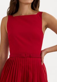 Rochie roșie cu decolteu pătrat și fustă plisată. Prezintă un corset ajustat și o curea asortată în talie. Textură uniformă, culoare vibrantă.