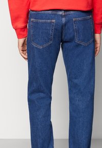 Jeans de mezclilla azul oscuro con corte recto, que cuentan con dos bolsillos traseros y costuras en contraste. La textura está ligeramente descolorida.