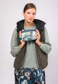 Gilet imbottito verde oliva sopra un maglione leggera verde, che tiene in mano una borsa cosmetica a fantasia con dettagli floreali blu e gialli e chiusura con zip.