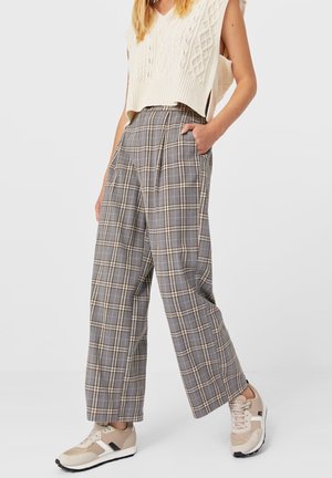 Kobieta ubrana w szare, szerokie spodnie w kratę, kremową dzianinową kamizelkę bez rękawów z bocznymi rozcięciami oraz beżowe trampki, stojąca z jedną ręką w kieszeni.