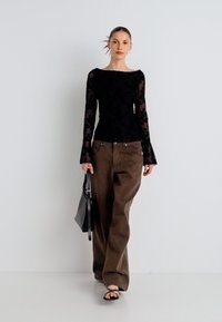 Črna bluzna s čipko, z dolgimi rokavi in štirikotnim izrezom, kombinirana z širokimi rjavimi hlačami. Model drži črno torbico in nosi črne sandale z naramnicami.