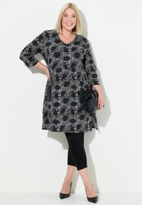 Schwarzes und weißes gemustertes Kleid mit Dreiviertelärmeln, einem V-Ausschnitt, seitlichen Taschen, kombiniert mit schwarzen Leggings und hohen Schuhen.