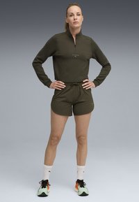 Olive roheline sporditopp poole tõmblukuga, millele on sobitatud sobivad lühikesed püksid. Omab lõdva istuvust, nööriga vöökohta ja kerget kangast.