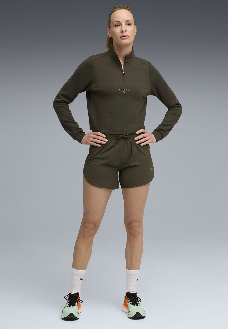 Olive roheline sporditopp poole tõmblukuga, millele on sobitatud sobivad lühikesed püksid. Omab lõdva istuvust, nööriga vöökohta ja kerget kangast.