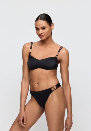 Vrouw met donker haar in een zwart bikinisetje met gouden accenten, staand tegen een effen lichtgrijze achtergrond, zacht glimlachend naar de camera.