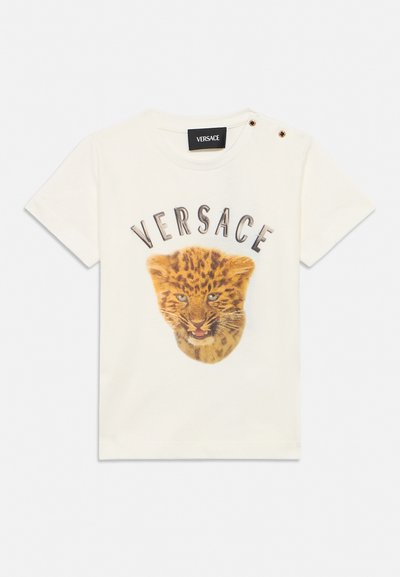 T-shirt crème à manches courtes avec le texte "VERSACE" au-dessus d'un visage détaillé de léopardon, avec deux boutons dorés sur l'épaule gauche.