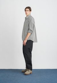 Camisa a rayas blancas y negras con un bolsillo en el pecho, combinada con pantalones negros y calzado marrón claro sobre una alfombra azul.