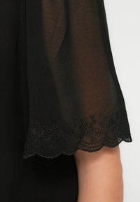 Μανίκι από μαύρο διαφανές ύφασμα με κυματιστή άκρη και κεντήματα με λουλούδια, που καλύπτει μερικώς έναν ανοιχτόχρωμο βραχίονα με φόντο λευκό.