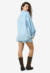 Chemise en denim bleu clair avec des rayures blanches verticales, coupe oversize, manches longues et pli au dos ; assortie à un short correspondant et des bottes marron jusqu'au genou.