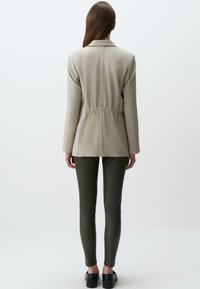 Hellbeige Jacke mit lässigem Schnitt, tailliert, kombiniert mit dunkelgrünen schmalen Hosen und schwarzen Schuhen. Glatte Stofftextur.