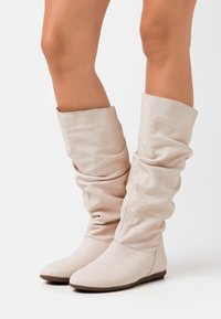 Botas de cuero beige suave, hasta la rodilla y con un diseño relajado, con una textura lisa y punta redondeada, que cuentan con una suela plana y un detalle de costura minimalista.