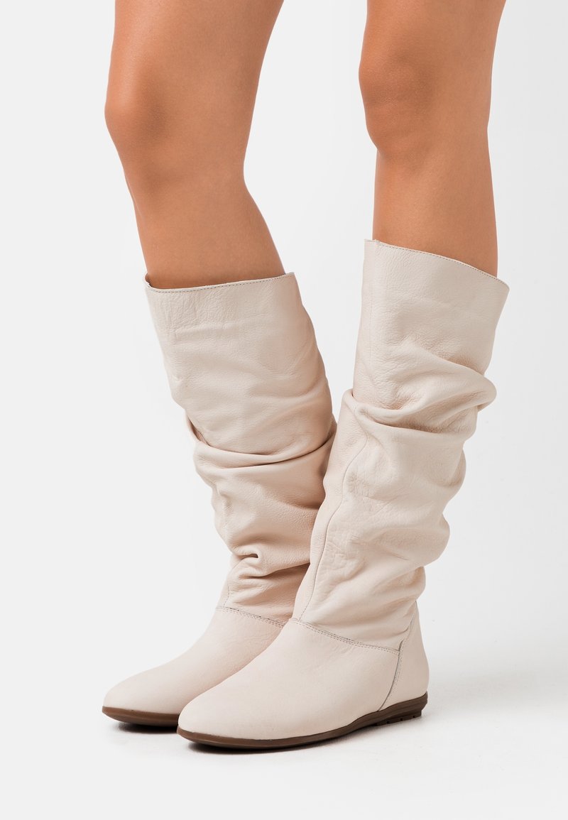 Botas de cuero beige suave, hasta la rodilla y con un diseño relajado, con una textura lisa y punta redondeada, que cuentan con una suela plana y un detalle de costura minimalista.