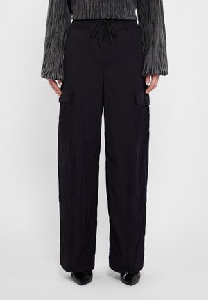 Schwarze Cargo-Hose mit weitem Bein, Kordelzug in der Taille, Reißverschlüssen am Saum und Klappentaschen, kombiniert mit einem grauen, strukturierten Langarmshirt und schwarzen spitzen Schuhen.