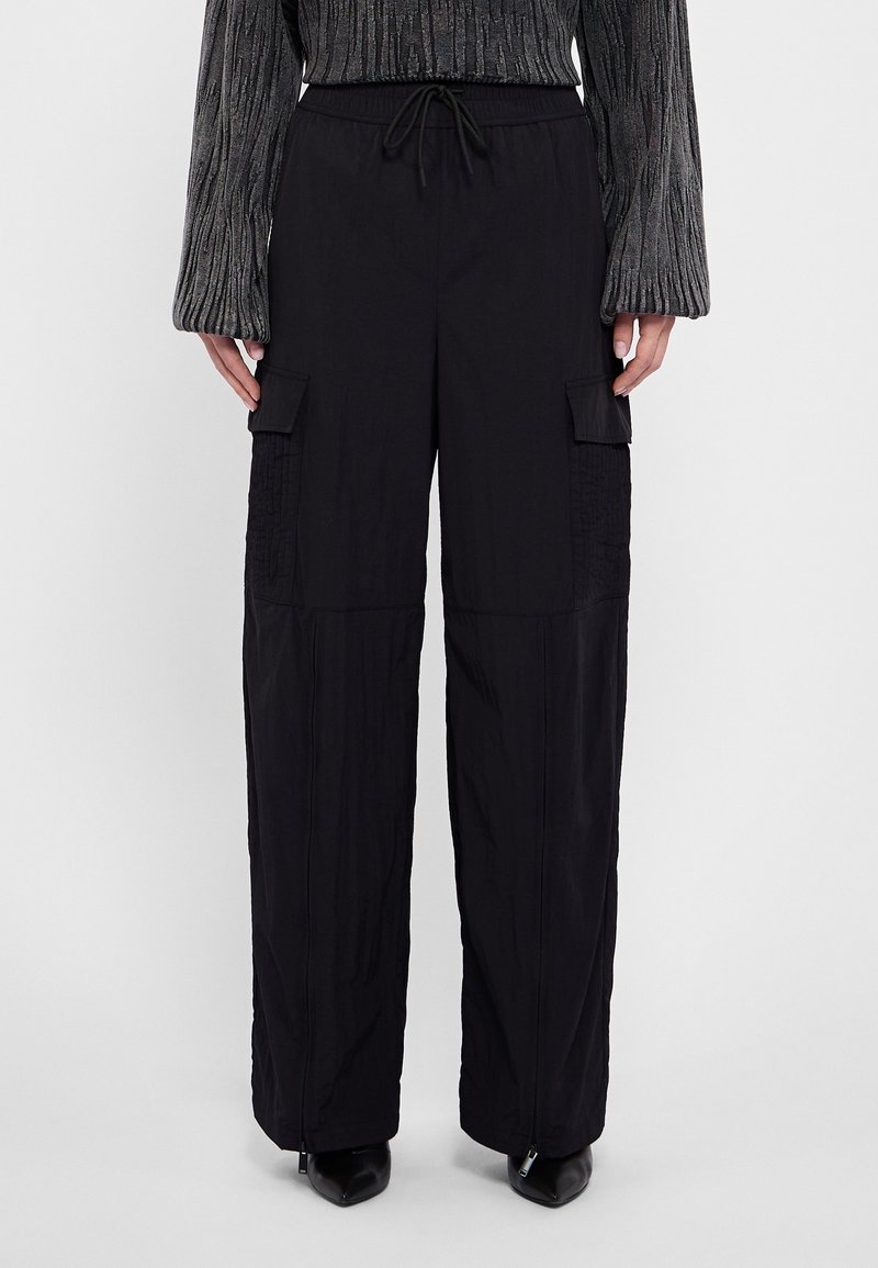 Pantalon cargo noir à jambes larges avec taille à cordon de serrage, ourlets zippés et poches à rabat, porté avec un haut gris texturé à manches longues et des chaussures noires à bout pointu.