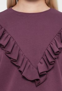 Sweatshirt violet avec un large volan en forme de V le long de l'encolure, fabriqué en coton doux avec une texture lisse.