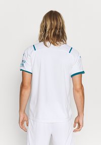 Bílý atletický dres s tyrkysovými akcenty na límci a rukávech, vyrobený z texturované tkaniny a se rovnými liniemi. Viditelné logo na rameni.