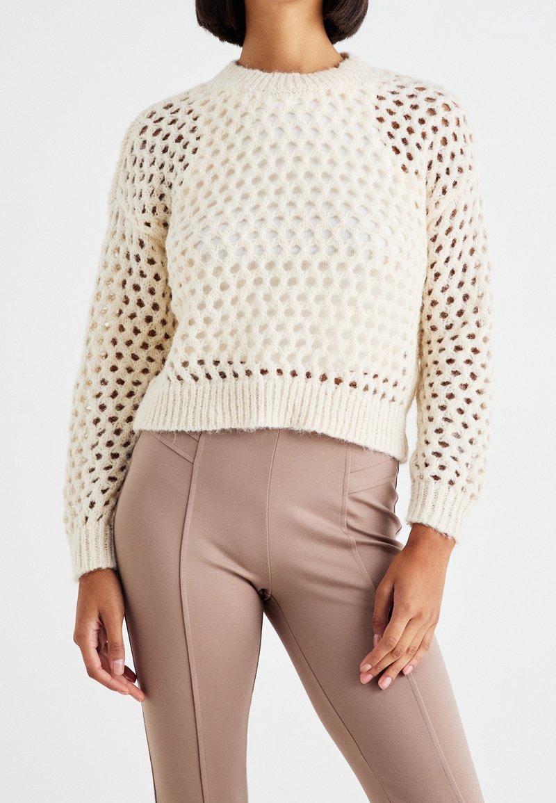 Femme portant un pull en maille ouverte de couleur crème à manches longues et un pantalon taupe taille haute sur fond blanc.