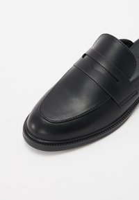 Black leather loafers with a rounded toe, featuring a decorative strap across the front and a textured outsole for grip.