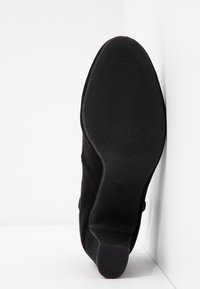 Semelle de chaussure en daim noir avec un fin motif ondulé et un talon bloc, positionnée verticalement contre un mur d’angle blanc, taille 37.