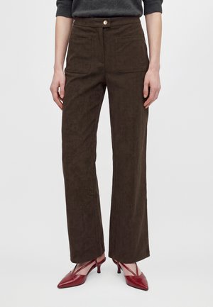 Un mannequin porte un pantalon marron foncé à jambes larges, doté de grandes poches à l’avant, et des escarpins rouges à bout pointu, sur un fond uni.