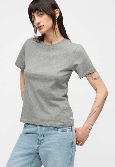 Γυναίκα με ίσια σκούρα μαλλιά, που φοράει γκρι μπλουζάκι και ανοιχτό μπλε τζιν, δείχνοντας τα τατουάζ στο δεξί της χέρι μπροστά σε απλό φόντο.