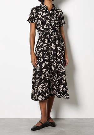 Vrouw draagt een zwarte midi-jurk met beige bladpatronen, korte mouwen, elastische taille en zwarte platte schoenen, staand op een grijze vloer.