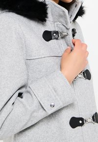 Šedý vlněný kabát s černým kožešinovým límcem, stříbrnými kovovými prvky a uzávěry na knoflíky. Obsahuje vysoký límec a zapínání na knoflíky na manžetách.