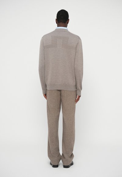 Άνδρας που κοιτάζει προς τα πίσω, φορώντας μπεζ πλεκτό πουλόβερ με γεωμετρικό σχέδιο και αντίστοιχες φαρδιές παντελόνες, όρθιος μπροστά σε λευκό φόντο.