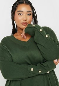 Green ribbed knit sweater with a V-neck and long sleeves, featuring gold button accents on the cuffs. Soft texture and relaxed fit.