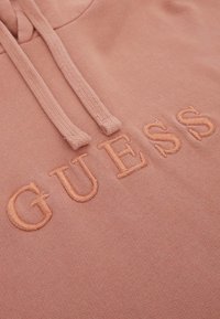 Rožinė sportinė striukė su žodžiu "GUESS" pakeltomis, siuvinėtomis raidėmis. Įtraukta kapa su reguliuojamais raišteliais ir minkšta audinio tekstūra.
