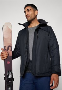 Svart isolert skijakke med glidelås og trykknapper, slitt over en grå skjorte. Personen holder en ski med rosa detaljer og bindinger.