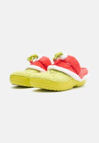 Green plush slippers with red fluffy tops, white trim, and decorative hands holding a red ornament. Textured yellow soles for grip.
