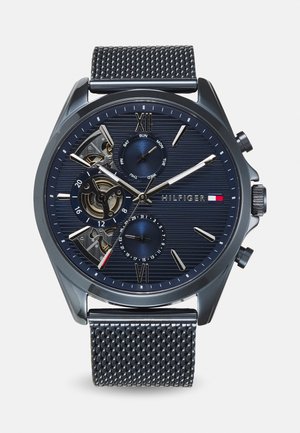 Pánske chronografické hodinky s modrým textúrovaným ciferníkom, striebornými hodinovými indexmi, odhalenými prevodmi a kovovým sieťovým remienkom. Medzi funkcie patrí indikátor dátumu a dňa.