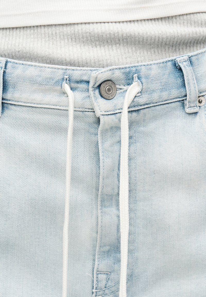 Light blue denim jeans with a metal button and white drawstring laces tied through belt loops over a ribbed white shirt.