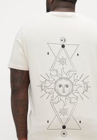 Kremna majica s kratkimi rokavi, ki ima na hrbtu črne geometrične in nebesne ilustracije, vključno z dizajnom sonca in lune.