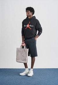 Μαύρο φούτερ με κόκκινο λογότυπο "Air Jordan", γκρι τσάντα, σκούρο σορτς και λευκά αθλητικά παπούτσια με πολύχρωμες λεπτομέρειες, τοποθετημένα σε απλό φόντο.