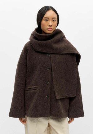 Femme aux cheveux noirs portant une veste en laine marron foncé oversize boutonnée et une écharpe en laine assortie sur un pantalon beige clair.