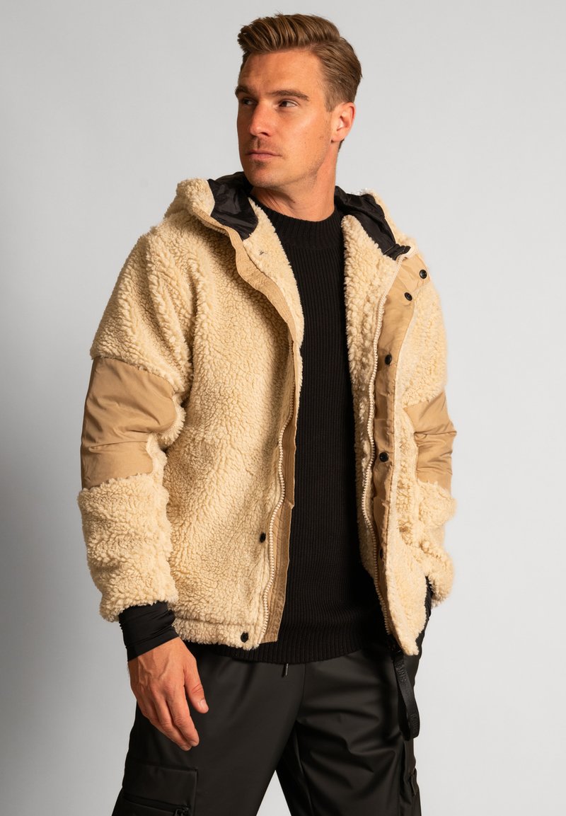 Giacca sherpa beige con maniche in tessuto marrone. Presenta un cappuccio, bottoni a pressione e un esterno testurizzato. Indossata sopra un maglione in maglia nera.