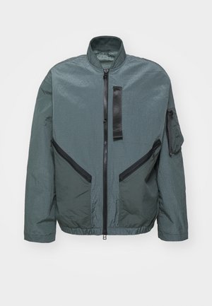 Green, lightweight jacket with a textured finish. Features a high collar, two front zip pockets, and an additional arm pocket with a flap.