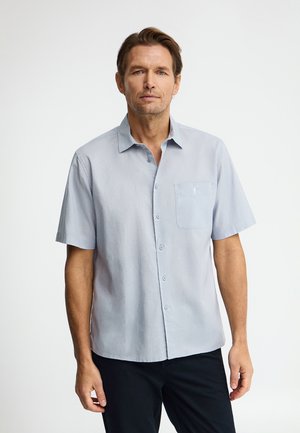 Homme portant une chemise bleu clair à manches courtes avec une poche poitrine, debout devant un fond blanc uni.