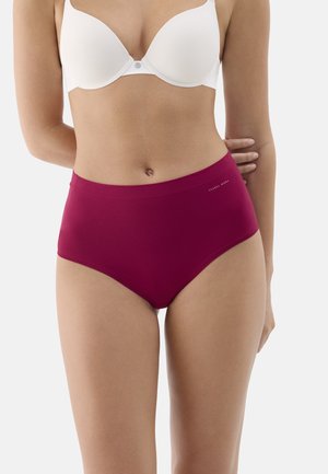 Mujer con sujetador blanco y bragas de tiro alto de color rosa oscuro, de pie sobre un fondo claro y liso.