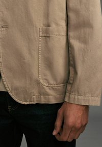 Veste beige clair avec une poche poitrine unique et des coutures surpiquées, fabriquée en tissu durable, portée sur un jean noir avec un poignet visible.