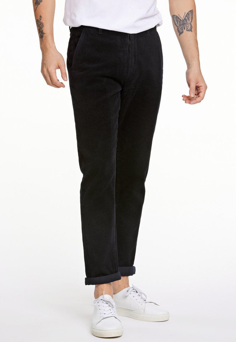Pantalones de pana negros con corte recto, que cuentan con bolsillos y dobladillos enrollados, combinados con zapatillas blancas. La textura es acanalada.